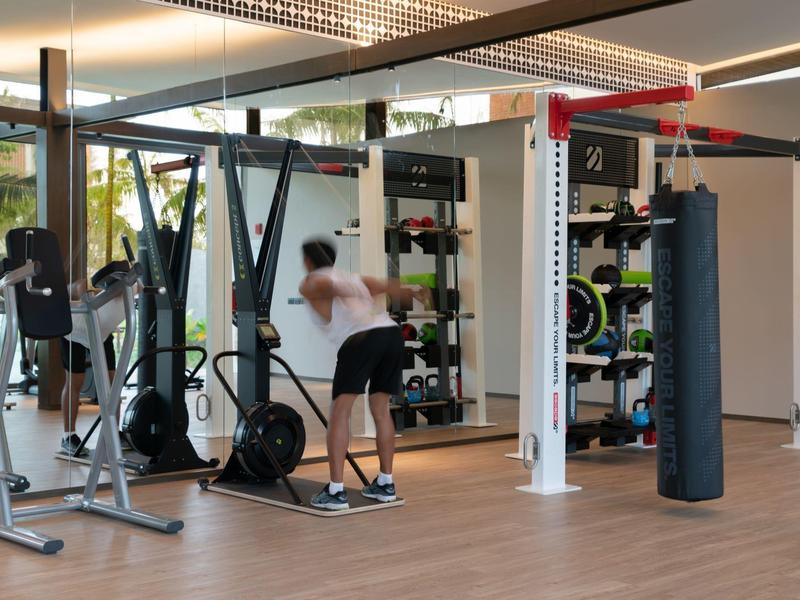 Gimnasio con máquinas y una persona levantando pesas, ventanas grandes y suelo de madera