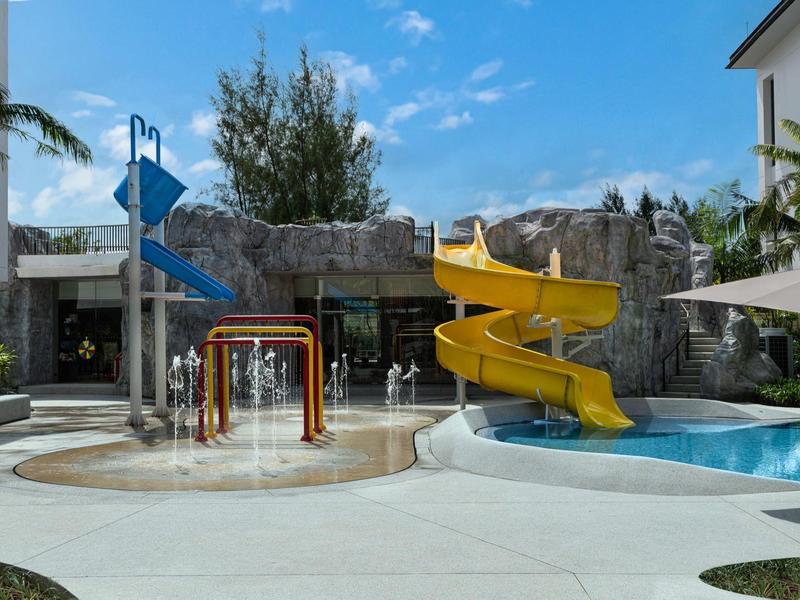 Piscina exterior con tobogán de agua amarillo, área de juegos acuáticos pequeña y sombrillas en un día soleado.