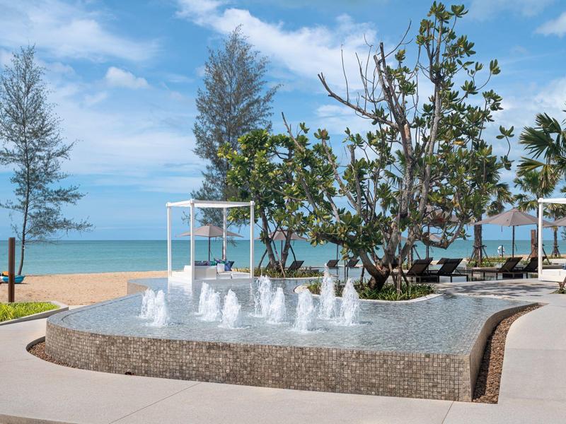Fuente moderna en la playa con árboles y cielo azul al fondo.