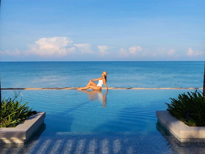 Mujer en traje de baño blanco disfruta de piscina infinita con vista al mar tranquilo y cielo azul.