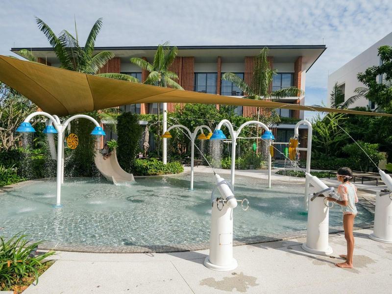 Niños juegan en una piscina infantil con fuentes y toldos frente a un hotel.