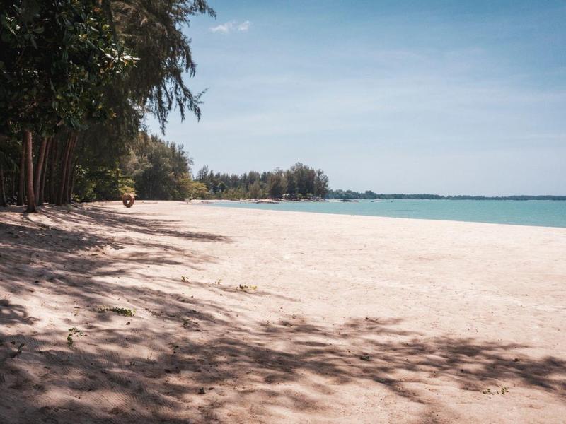 Opuszczona piaszczysta plaża z drzewami po lewej stronie i spokojnym niebieskim morzem pod jasnym niebem.