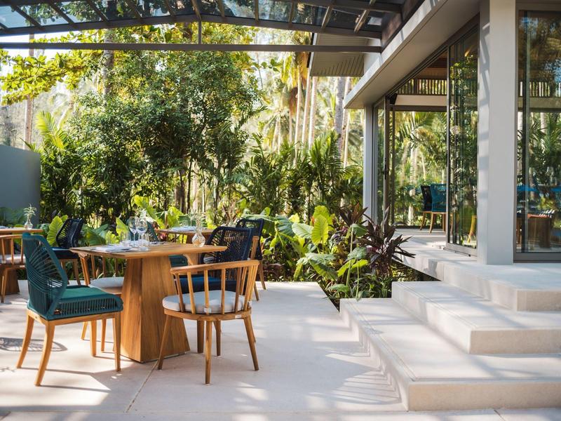 Słoneczny taras hotelowy z drewnianym stołem, mieszanymi krzesłami i bujną zielenią roślin.