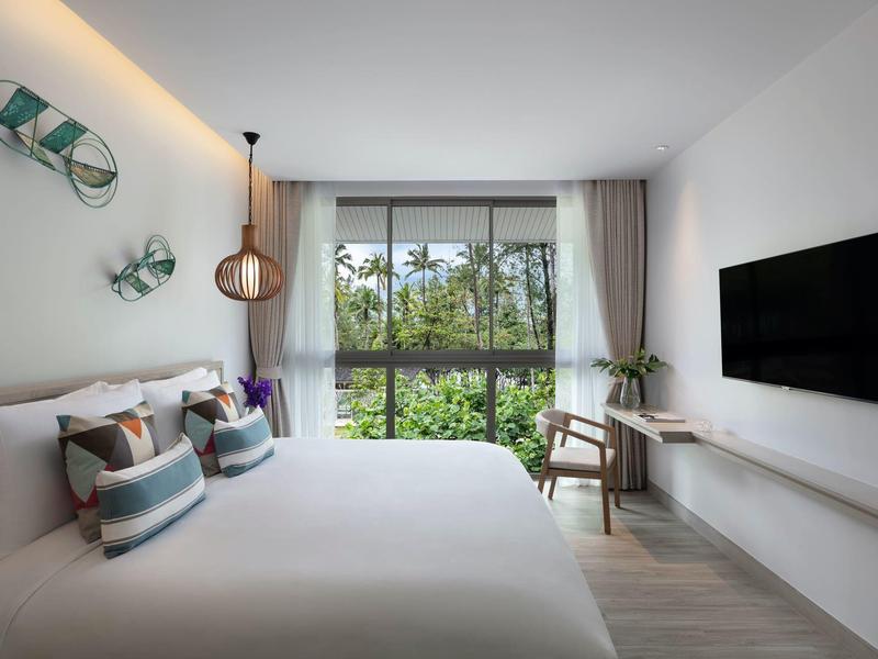 Nowoczesny pokój hotelowy z dużym łóżkiem, telewizorem na ścianie i widokiem na zieloną przyrodę.