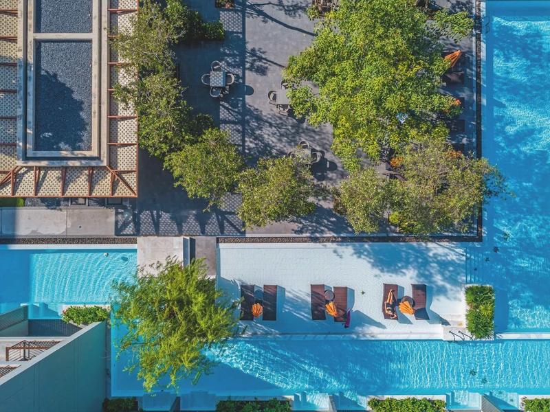 Pohled z ptačí perspektivy na bazénovou plochu s stromy a lehátky vedle hotelu.