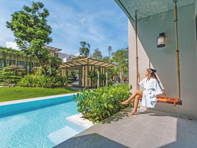Žena na houpačce vedle bazénu s výhledem na zahradu a pergolu za slunečného dne.