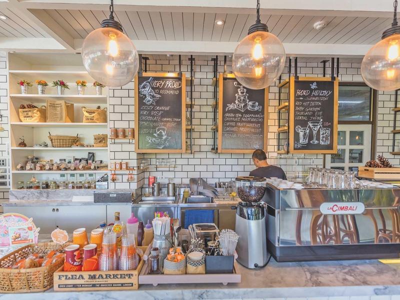 Světla pekárna s dobře zásobenými regály, kávovarem a mužem za pultem.