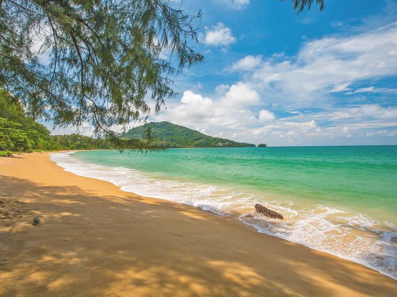 Klidná písečná pláž se zelenou vodou a zalesněným kopcem pod slunečnou oblohou.