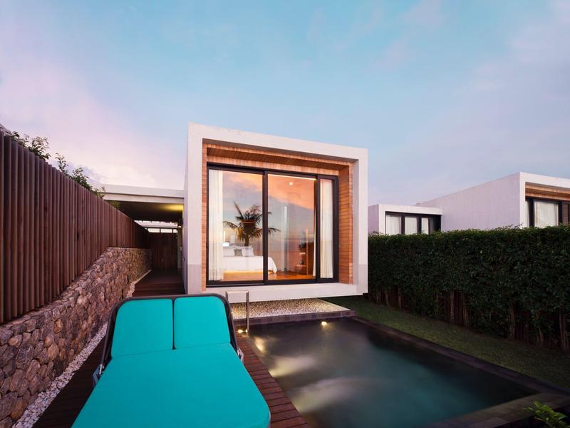 Moderní bazénová plocha s lehátkem a skleněným hotelem za západu slunce.