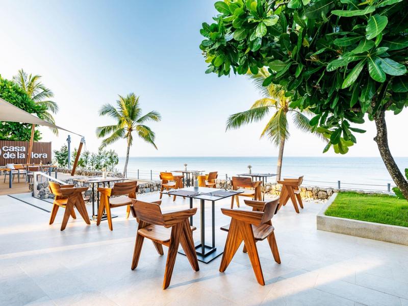 Otevřená sezení s dřevěnými stoly a židlemi na pláži s palmami a výhledem na moře.