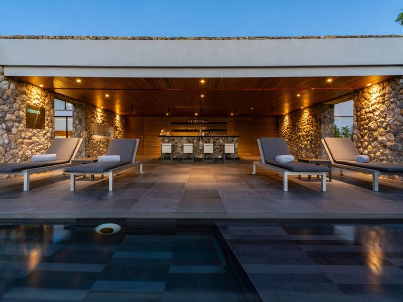 Moderní bazénová oblast s lehátky a osvětleným lounge pod dřevěným obložením.