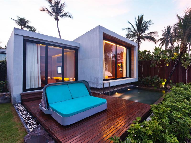 Moderní rekreační dům s velkým oknem a modrou pohovkou na dřevěné terase obklopené palmami.