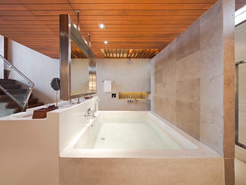 Moderní vana v světlé koupelně s dřevěným stropem a schody v pozadí.