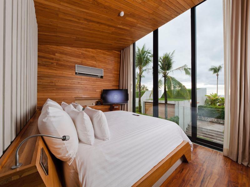 Moderní hotelový pokoj s dřevěnými stěnami, velkou postelí a výhledem na palmy přes celoplošná okna.