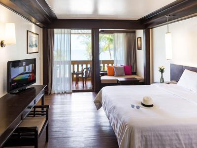 Helle, moderne Schlafzimmer mit großem Bett, Fernseher, Sofa und Balkon mit Blick ins Grüne.