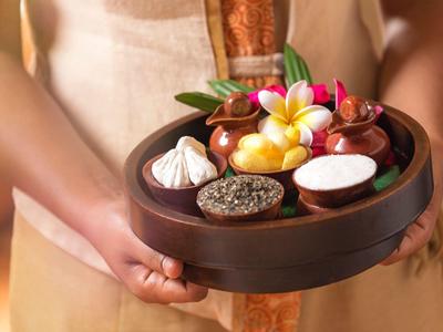 Person hält braunes Holztablett mit kleinen Schalen, Blumen und Tropfen für Spa-Behandlung.