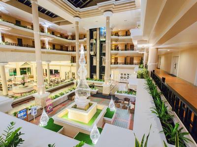 Großes, helles Atrium mit Springbrunnen, grünen Pflanzen und mehrstöckigen Gängen in warmen Farben.