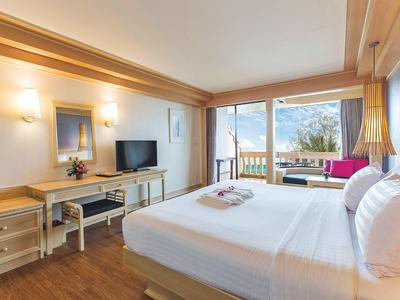 Lichte hotelkamer met groot bed, houten vloer, tv en balkon met uitzicht op bomen.