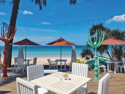 Strandrestaurant mit weißen Tischen, Stühlen, rotbraunen Sonnenschirmen und blauem Meer im Hintergrund.