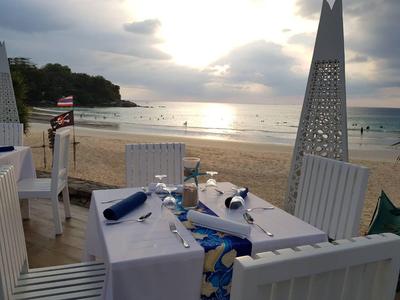 Ein gedeckter Tisch mit weißen Stühlen am Strand, Sonnenuntergang und ruhiges Meer im Hintergrund.