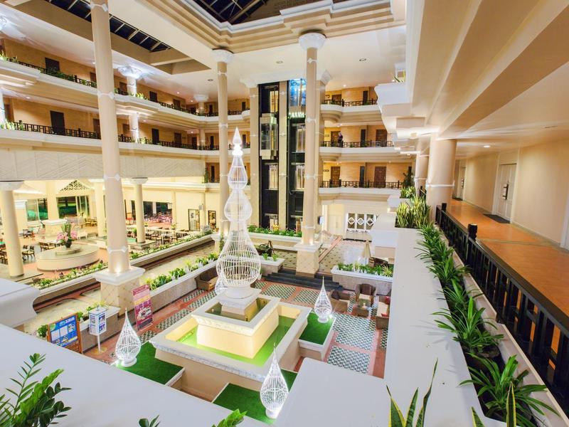 Großes, helles Atrium mit Springbrunnen, grünen Pflanzen und mehrstöckigen Gängen in warmen Farben.