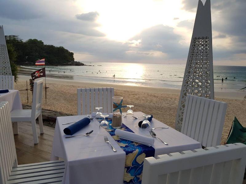 Ein gedeckter Tisch mit weißen Stühlen am Strand, Sonnenuntergang und ruhiges Meer im Hintergrund.