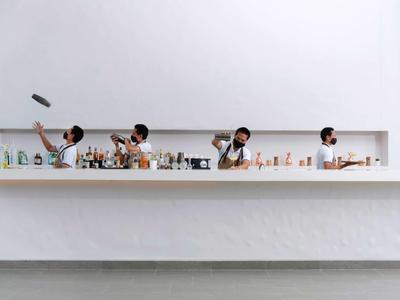 Barman mezcla cócteles detrás de una larga barra blanca en un espacio minimalista.