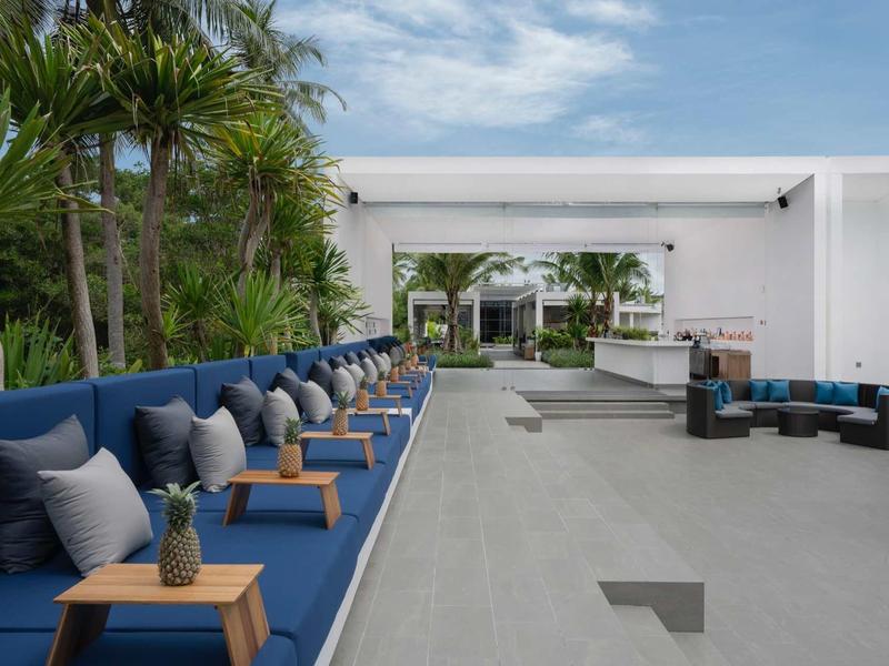 Área exterior moderna con bancos azules y pequeñas mesas de madera junto a la piscina del hotel.