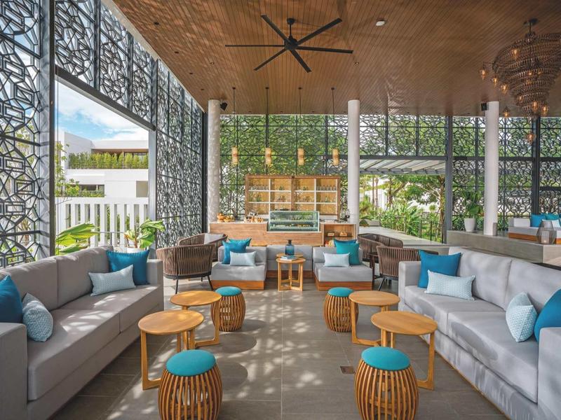 Área de salón moderna con sofás grises, cojines azules y bancos de madera en un hotel.