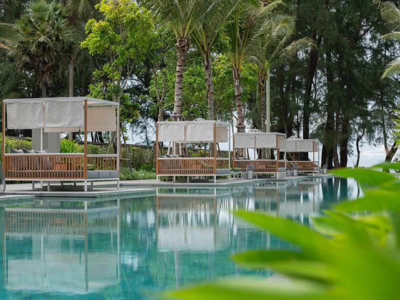 Una zona tranquila de piscina con camas balinesas y vegetación verde al fondo en un día soleado.