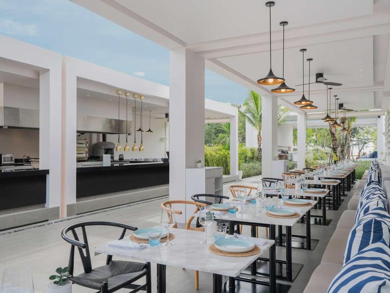 Área exterior moderna de un hotel con mesas, sillas y cojines coloridos en un banco largo.