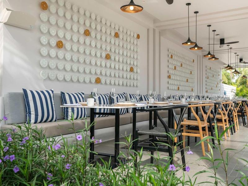 Área exterior moderna de un hotel con paredes blancas, bancos cómodos y mesas de madera.