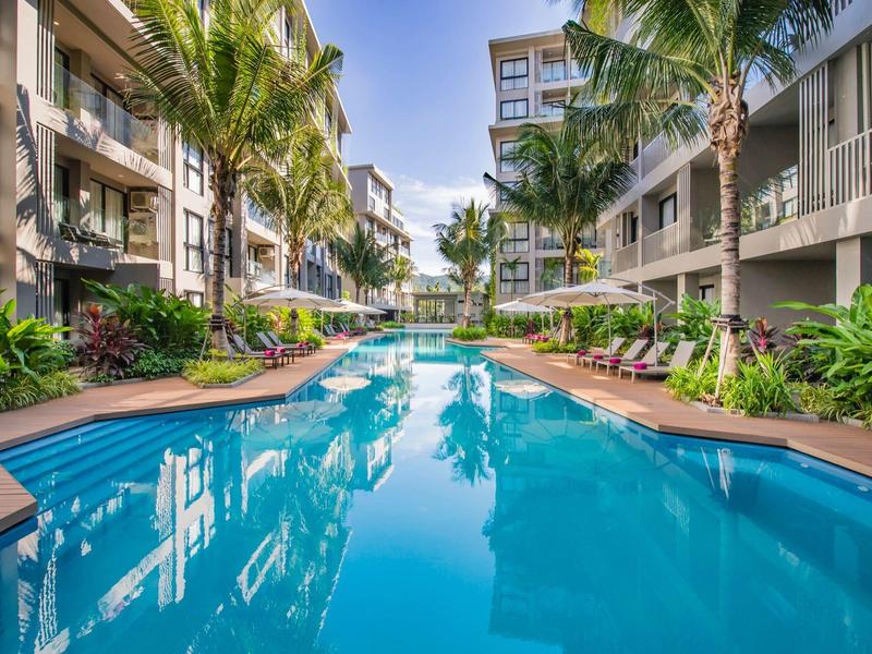 Modern hotel pool between two-story buildings with palm trees and lounge chairs.