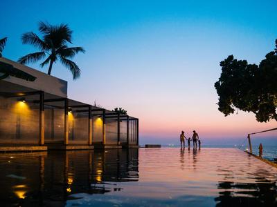 Abendliche Terrasse mit Pool, Palmen und Menschen vor einem bunten Himmel am Meer.