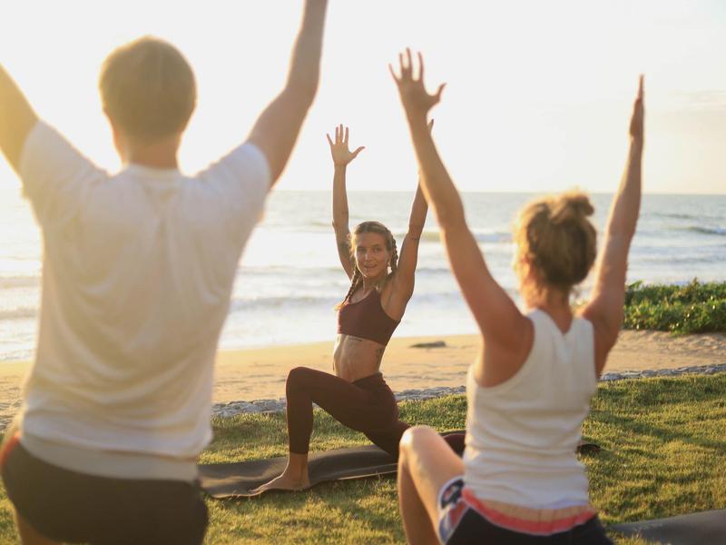 Drei Personen praktizieren Yoga im Freien am Strand bei Sonnenuntergang.