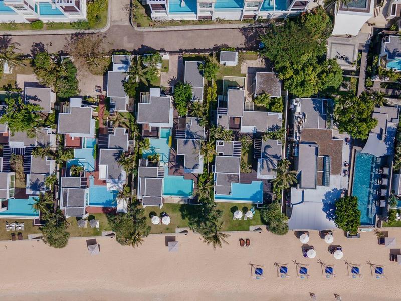 Luftaufnahme einer Strandhotelanlage mit Pools und Sonnenschirmen am Sandstrand.