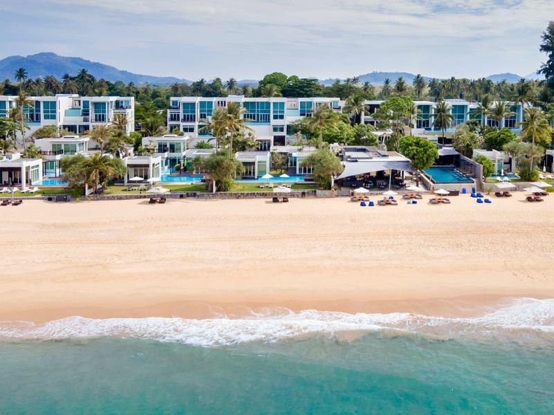 Moderne weiße Strandvillen mit blauen Fenstern, Palmen, Sandstrand und ruhigem türkisem Meer.