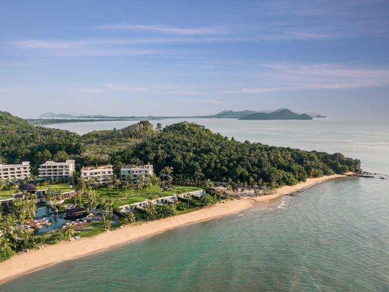 Complesso alberghiero sulla spiaggia con colline verdi e mare calmo sotto un cielo blu.