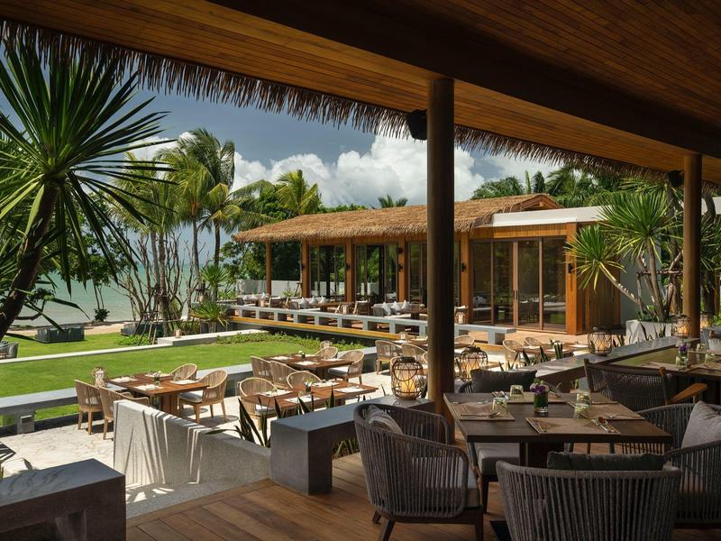 Area ristorante all'aperto con mobili in legno, vista su piante tropicali e mare al sole.