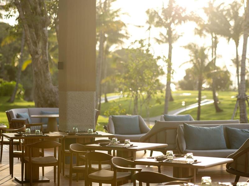 Terrazza accogliente con tavoli e sedie in legno, circondata da palme e luce solare.
