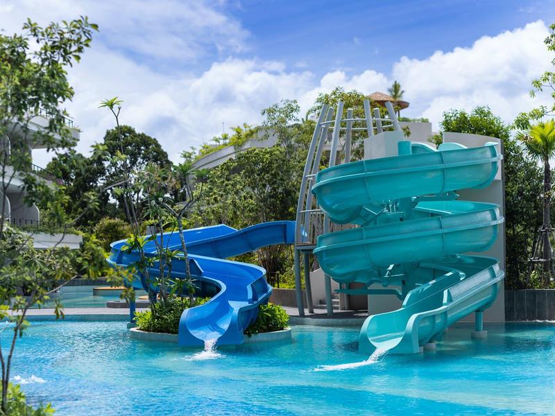 Una piscina all'aperto con due scivoli d'acqua blu circondata da alberi e cielo azzurro.