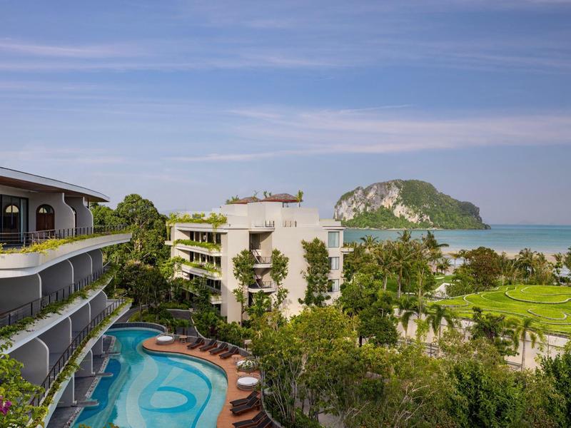 Complesso alberghiero con piscina, vegetazione rigogliosa e vista sul mare e una formazione rocciosa sullo sfondo.