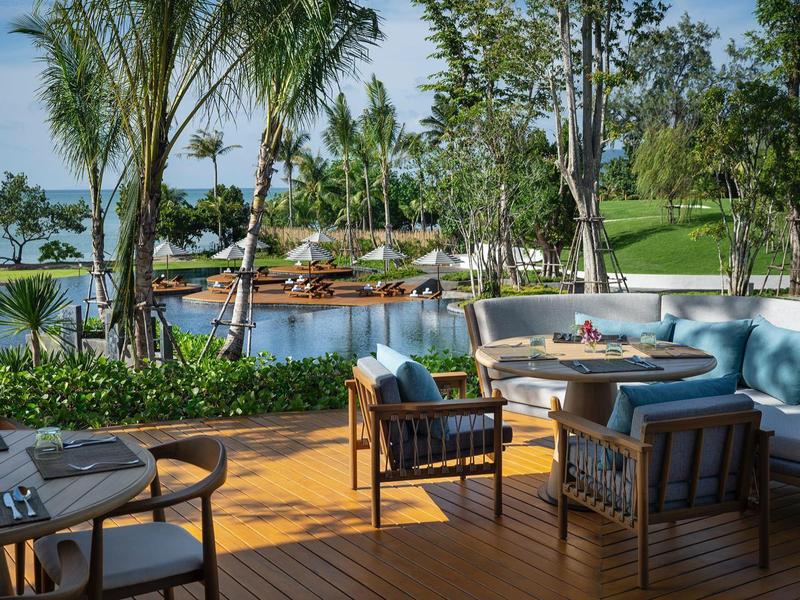 Area esterna con poltrone e divani confortevoli su terrazza in legno accanto a piscina e giardino tropicale.