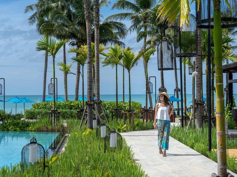 Žena kráčí po cestě lemované palmami vedle bazénu s výhledem na moře.