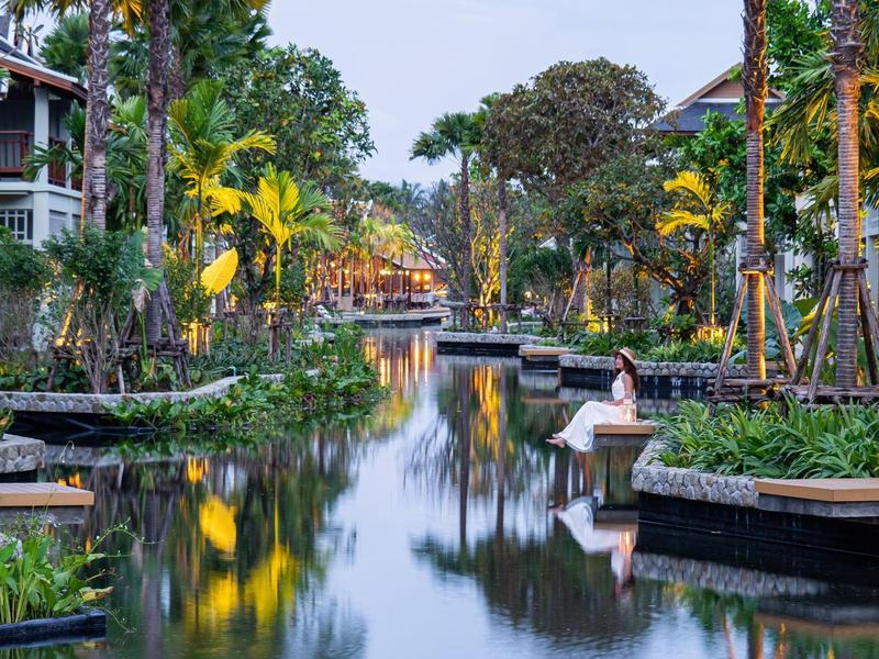 Vodní kanál s lavičkami a tropickou vegetací v hotelovém komplexu za soumraku.