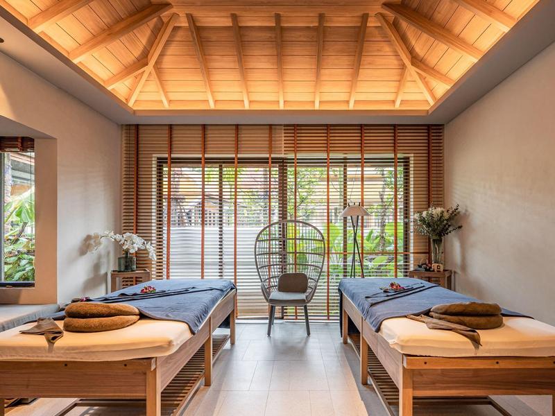 Světelná spa místnost se dvěma masážními lehátky, proutěnými křesly a dřevěným stropem s nepřímým osvětlením.