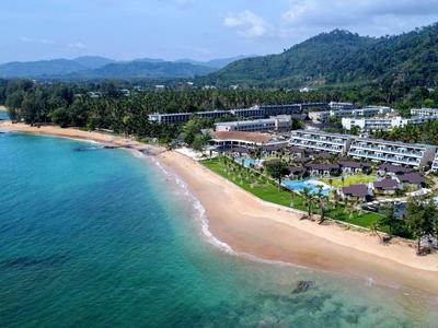 Panoramatický pohled na písečnou pláž s tyrkysovou vodou a resortem u zelených kopců.