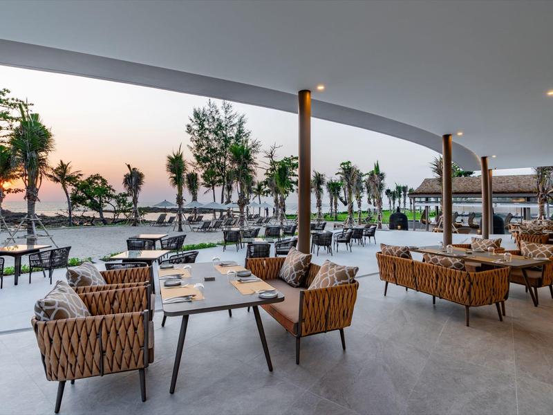 Otevřený hotelový prostor se sezením a výhledem na palmy a západ slunce na pláži.