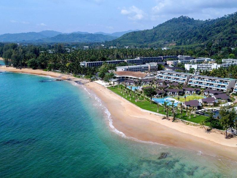 Strand mit türkisfarbenem Wasser und Hotels im Hintergrund vor bewaldeten Hügeln.