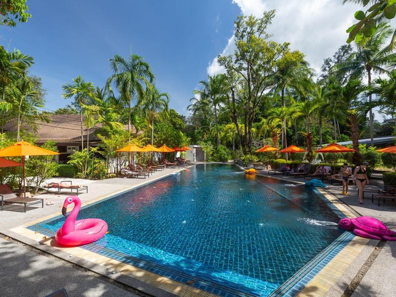 Grande piscina con piastrelle blu, gonfiabili fenicottero rosa e ombrelloni in un resort tropicale.